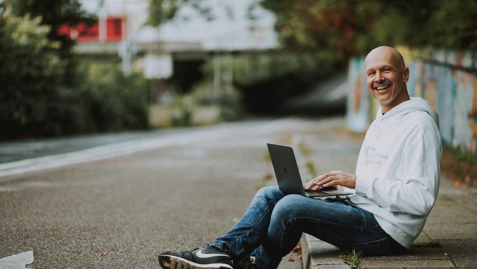 Webdesigner Paul Jackson sitzt am Bordstein mit Laptop und lächelt